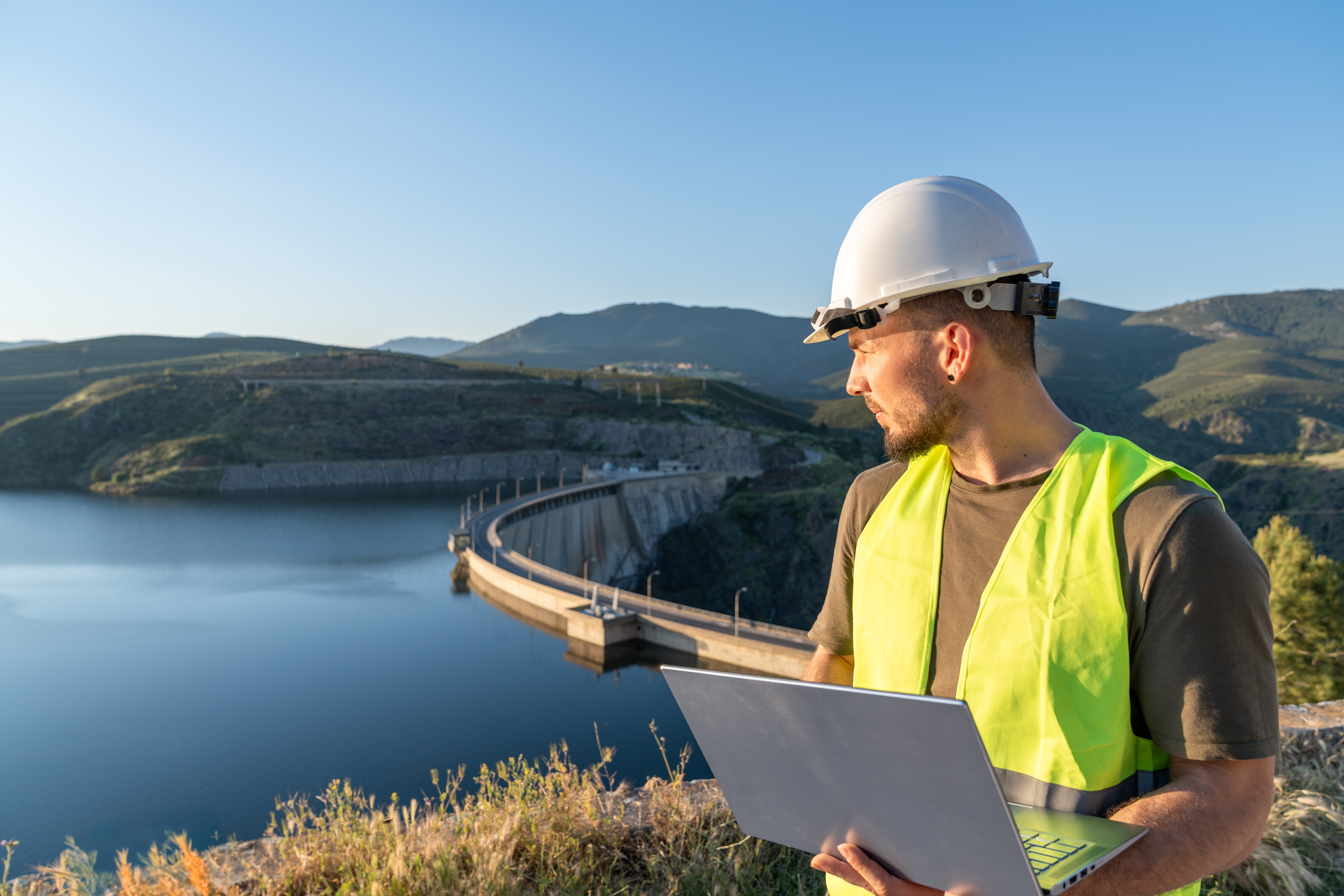 Engineer wearing a safety vest and helmet, holding a laptop while inspecting a reservoir dam, ensuring efficient water resource management